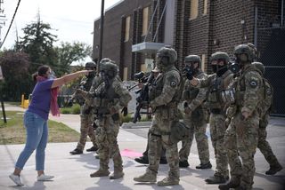 a person wearing a respirator pointing at a half dozen federal agents in full camo gear with weapons
