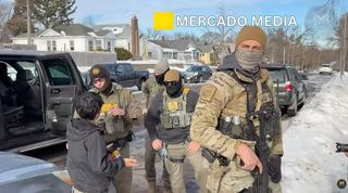 From left-to-right, a silver SUV is partially visible with the back rear door open all the way. Three masked agents stand talking to a young boy, who appears Latino, with a bag of chips in his hands. Edgar Vazquez is in the foreground facing the camera wearing a tan knit cap, a gray-green mask over the bottom half of his face, a dark grey hoodie under his camo fatigues holding his launcher. His ID patch EZ-17 is partially visible.