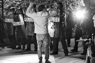 A person with hands in the air in front of a line of police with riot shields.