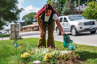 a woman with a watering can waters flowers along the road side