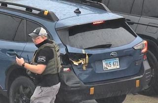 blue subaru plate 945 671 with taped rear bumper; a man in a baseball cap and body armor vest stands nearby