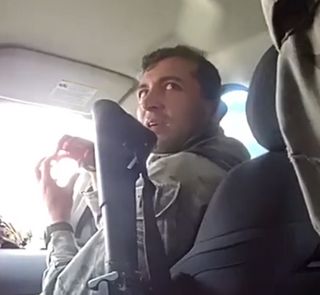 A man with dark hair sits in the front passenger seat of an SUV. The handle of a launcher is visible between the camera and man. He holds both hands up with fingertips touching, perhaps indicating a ball shape.