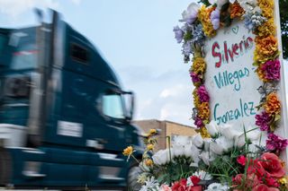 A sign adorned in colorful flowers reading Silverio Villegas Gonzalez at the memorial. A semi-truck is blurred in the background on the busy street.