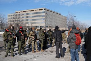 Over a dozen federal agents in a mixture of camo fatigues and dark green uniforms stand opposite roughly 7 protesters standing as one closest to the agents dressed in black points off to the right. One of the agents in a brown knit cap carrying a launcher is Edgar Vazquez.