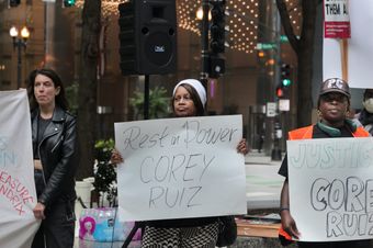 Three protesters in front of city buildings. A Black woman in the middle with a white cap is holding a white sign with “Rest in Power COREY RUIZ” scrawled on it in black marker.