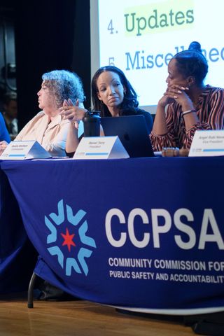 Close up image of three women sitting at a spotlighted table covered in a dark blue banner with the text "CCPSA" overlaid. Behind them is a large screen. A name placard for Sandra Wortham sits in front of her. She is looking at the person next to her with her eyebrows raised.