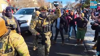 A chaotic scene showing residents in the background shouting at agents in the street. A white SUV is in the background. Several agents are partially visible while agent Edgar Vazquez dressed in camo fatigues, wearing a gray-green mask, and a tactical helmet holds a can of CS gas over his head, seemingly as a warning to the crowd.