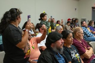 In a packed room, people sit and stand, many holding yellow flowers.