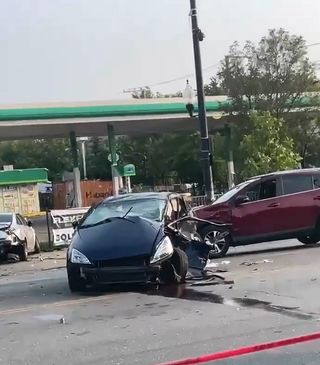 Scene of the crash with a black car severely damaged on the driver's side where it looks like the fleeing car t-boned it with two other damaged cars in front of a gas station.