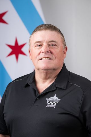 A white man with short-cropped hair in a black Chicago Police polo shirt with the Chicago flag in the background.