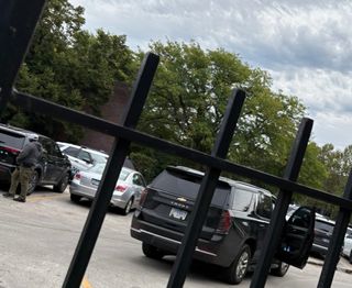 A man wearing a hoodie, a black mask and a white baseball cap stands next to a black SUV parked in a parking lot near a Chicago police cruiser behind a black metal fence. The SUV has an Illinois plate reading GC 12059.