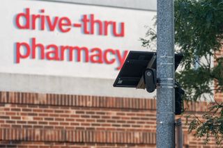 Black device that looks like a surveillance camera affixed to street pole in foreground; pharmacy sign in the background