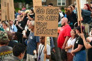 young person holding vigil candle and cardboard sign that reads "go back to Ohio you racist"