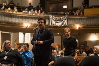 Chicago Alderman Byron Sigcho-Lopez speaks into a microphone as a crowd looks on and a younger man stands behind him. In the background, a banner reading "abolish ICE" was hung over a seating balcony.