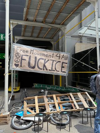 A homemade banner attached to scaffolding blocking a parking garage; it reads Free Movement 4 All fuck ice. There is also a pile of stuff in the driveway including bicycles, a wood palette and a ladder