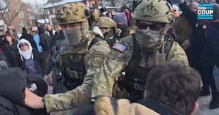 Two male Border Patrol agents dressed in military camouflage fatigues are close to the camera, shoving residents just in front of the camera. Both men are wearing ballistic helmets, their faces are hidden by masks. The one on the right is wearing dark sunglasses with a flag patch on his right arm. The man on the left wears an identification patch "EZ-17." "Police BORTAC" is partially visible on one vest.