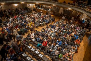 From above, a view of many rows of people seated in chairs inside a warmly lit auditorium.