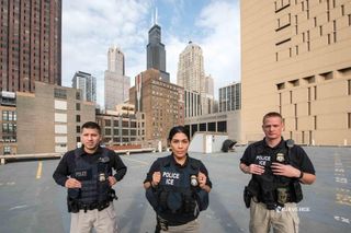 three federal agents in black shirts with dark vest and police badges pose on parking garage roof of downtown ICE building with skyscrapers seen in background