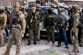 A group of border patrol aents stand around. Agent EZ-17 stands with his launcher pointed at the ground, wearing a mask and a multi-colored belt of various chemical and foam munitions. 

On the right, EZ-2 stands with a can of pepper spray. In the middle stands a shorter, older man with a tear gas canister dangling from his tactical vest.

Several other agents stand with them in front of a police vehicle.