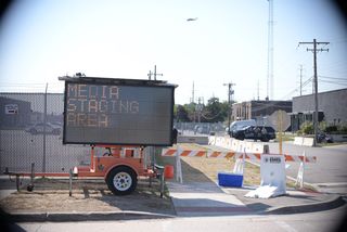 in the foreground, a mobile LED sign reading MEDIA STAGING AREA stationed in front of concrete barricades and traffic barriers. in the background, police vehicles and a helicopter.