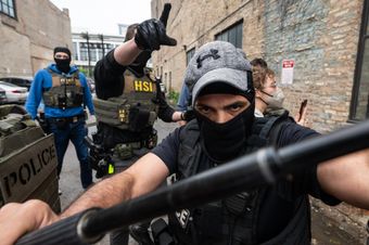 close up of masked ICE agent in an alley pushing back against viewer/photographer with a baton