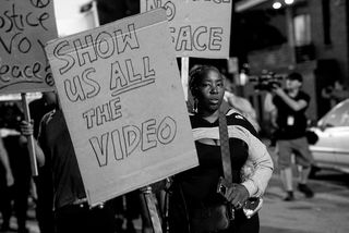 black and white photo of a protester holding a handmade cardboard sign reading SHOW US ALL THE VIDEO