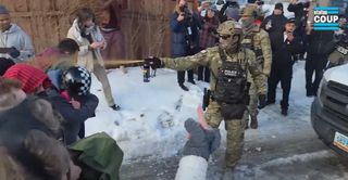 A masked border patrol agent wearing dark sunglasses stands in the middle of the road holding a large can of professional-grade pepper spray. A stream of spray hits several residents in the face as they stand approximately 6 feet from the agent. The agent has a tactical vest reading POLICE / BORTAC with an American flag patch on his right arm and helmet.