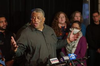 A Black man in a black shirt speaks to the press with supporters in the background.