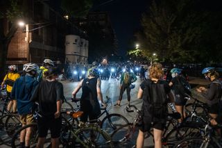 protesters on bikes at nighttime, viewed from behind, face off with police with bikes and bright lights in the distance