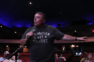 A man with a microphone in an auditorium. He is extending his arm while speaking and his shirt reads PATRIOTISM NOT POLITICS