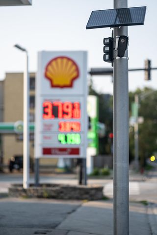 A Flock ALPR mounted to a utility pole with solar panels placed above it. A Shell gas sign is blurred in the background.