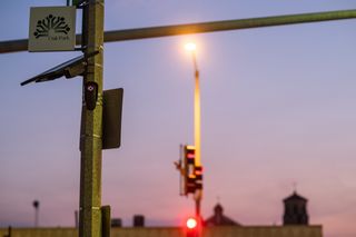 A Flock ALPR with light on the front illuminated and glowing red. It is affixed to a street pole and there is a sign for the village of Oak Park. Sky is purple/pink in the background.